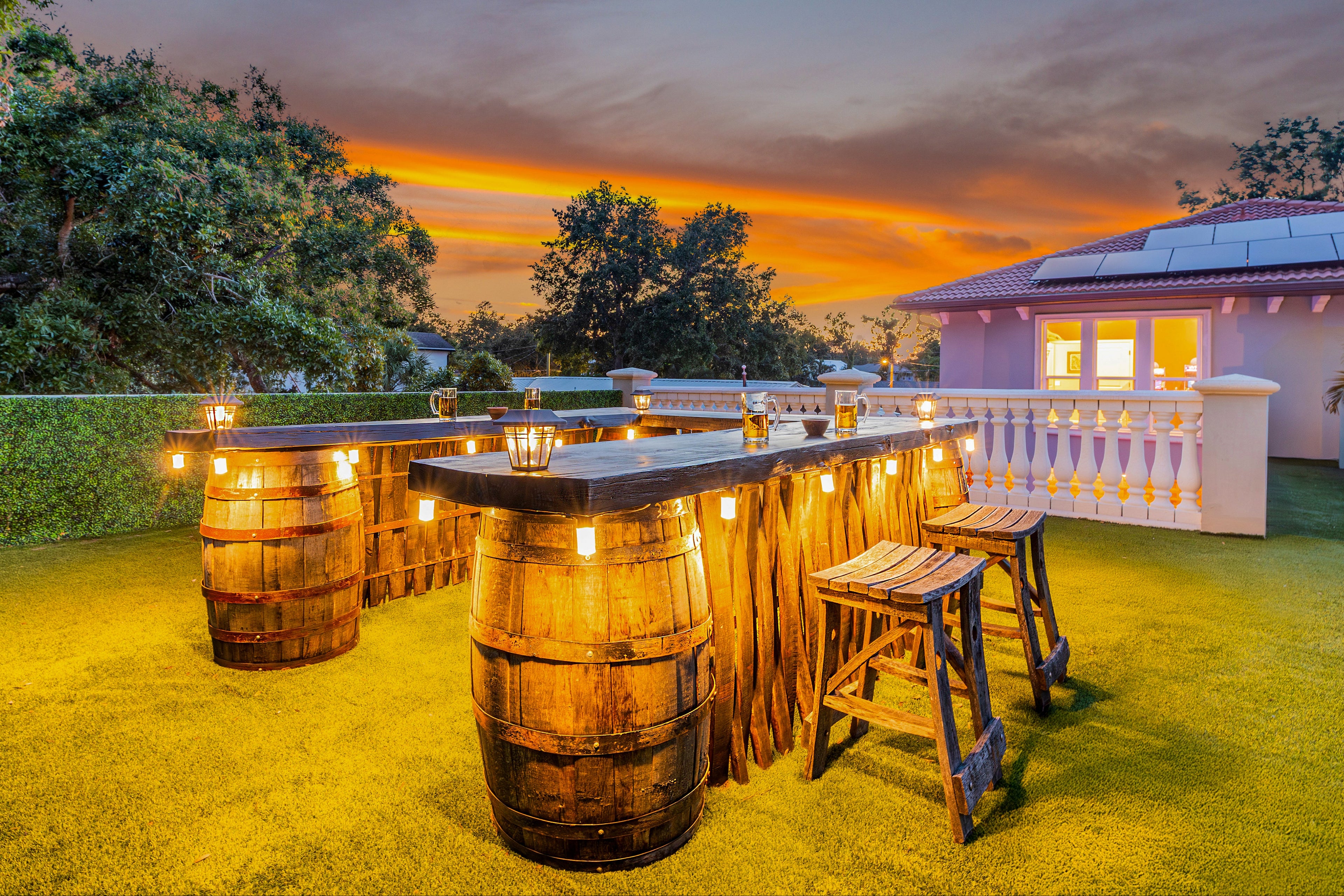 Outdoor bar setup with wooden barrels and stools against a sunset sky.