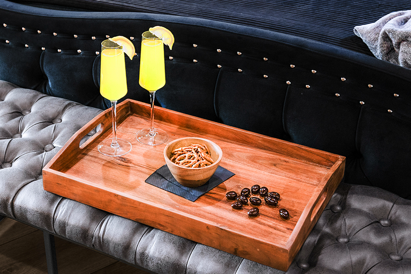 Wooden tray with mimosas and snacks on a tufted grey velvet bench in Gaspar's Quarters at Gaspar's Estate Tampa.